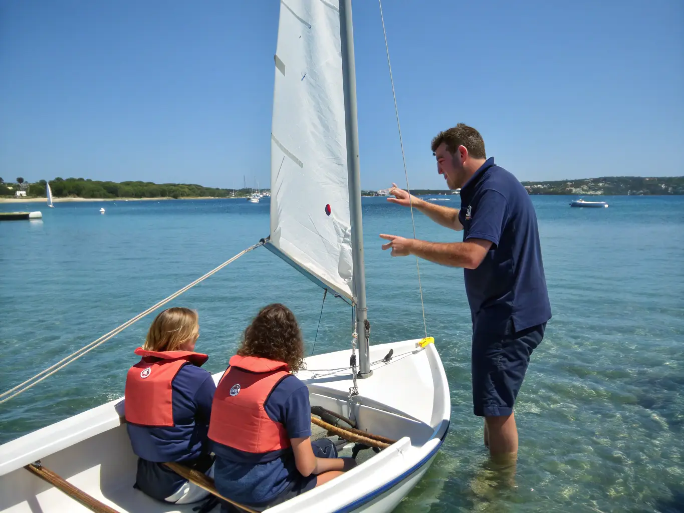A dynamic image of a sailing coach providing hands-on instruction to a student, demonstrating proper techniques and offering encouragement, with the sailing boat in motion on the water.